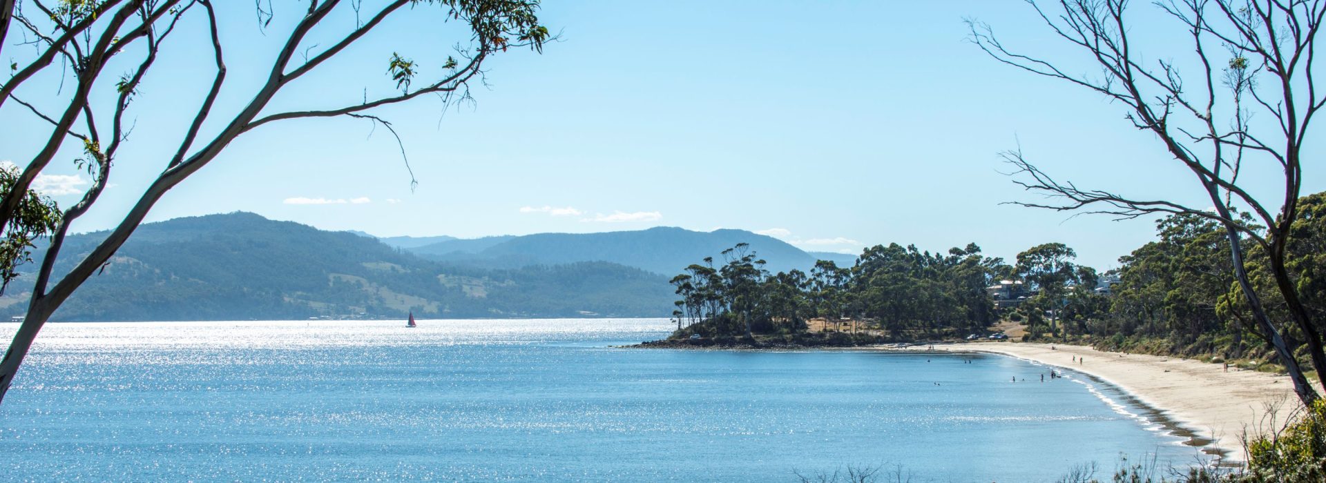 A calm beach with gentle waves, bordered by trees and hills under a clear blue sky. People walk along the sandy shore.