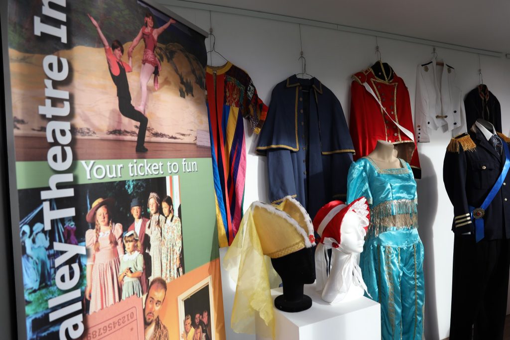 A display of colorful theatrical costumes and a mannequin head with hats, next to a banner reading Valley Theatre. Outfits include dresses, uniforms, and a blue satin ensemble on hangers against a white wall.