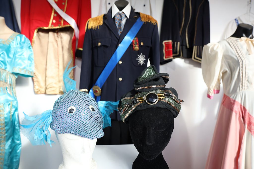Various theatrical costumes on mannequins, including a black suit with medals, colorful dresses, and two unique hats—one blue with fins and polka dots, the other dark with sequins and a large gem.
