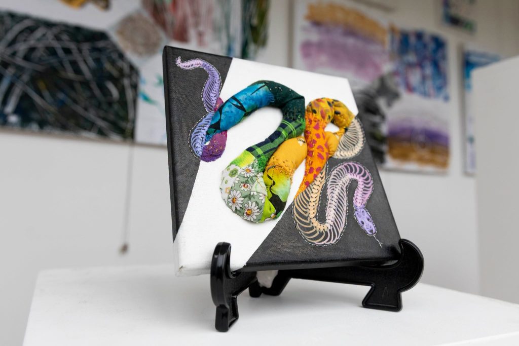 A small canvas on a black easel displays three colorful snake designs, one patchwork fabric and two painted, against a black-and-white background. Blurred abstract art hangs in the background.