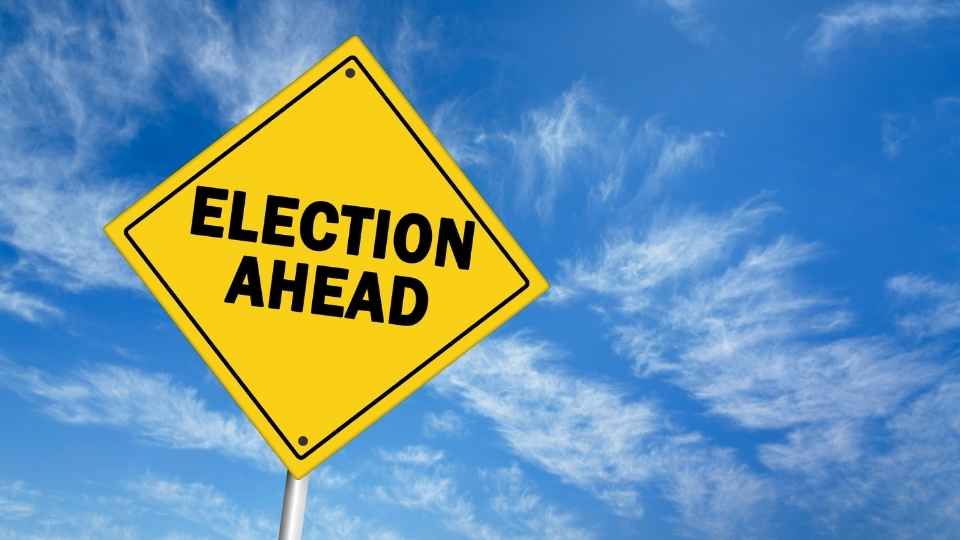 A yellow road sign with black text reads ELECTION AHEAD against a blue sky with wispy clouds.