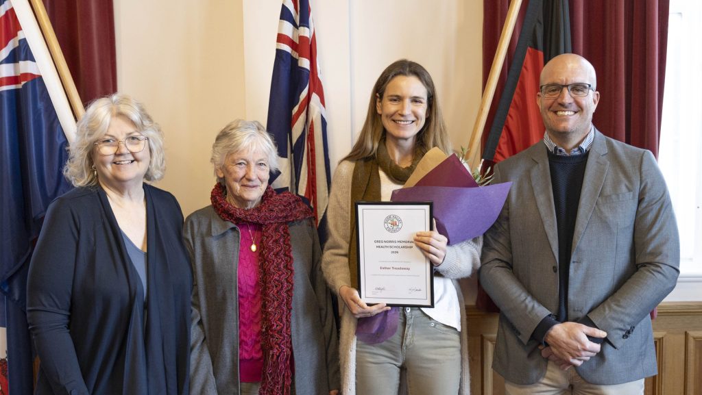 Four adults stand together indoors, smiling. The woman second from the right holds a certificate and folder. Flags and red curtains are visible in the background. The group is dressed in business casual attire.