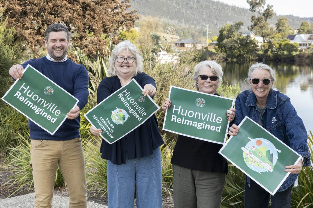 Four smiling adults stand outdoors by a river, holding green Huonville Reimagined signs, with trees, houses, and hills in the background.
