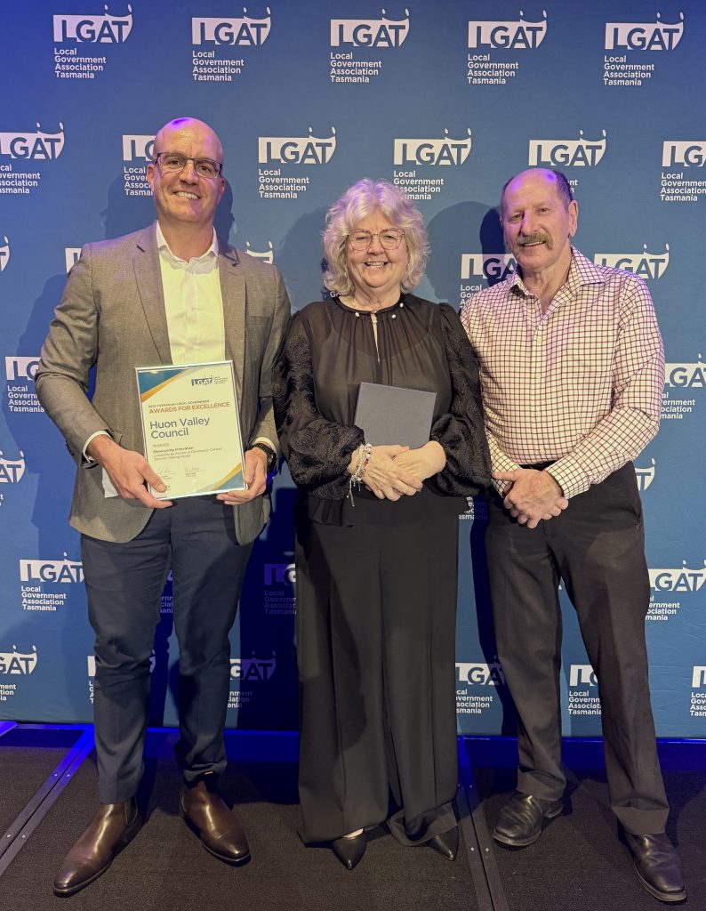Three people stand smiling in front of an LGAT backdrop. The person on the left holds a framed certificate labeled Huon Valley Council. The group is dressed in business and formal attire.