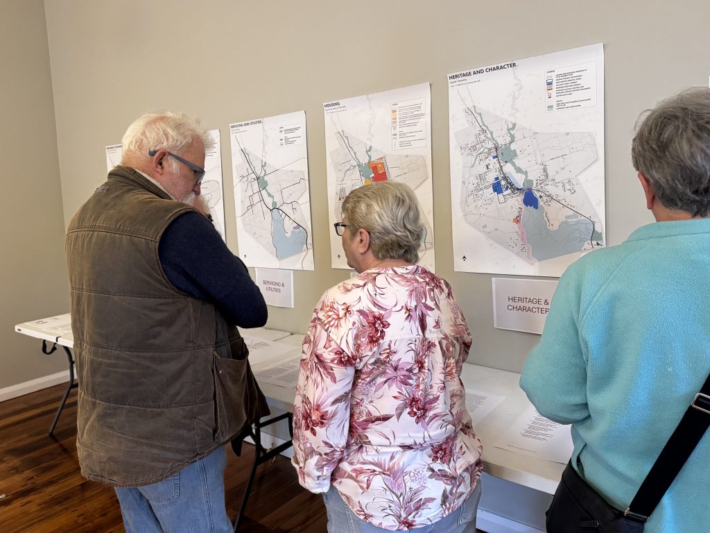 Three people stand at a table, viewing maps and documents displayed on the wall. The maps appear to show heritage and character areas. Two people are engaged in conversation, while one person looks at the maps.