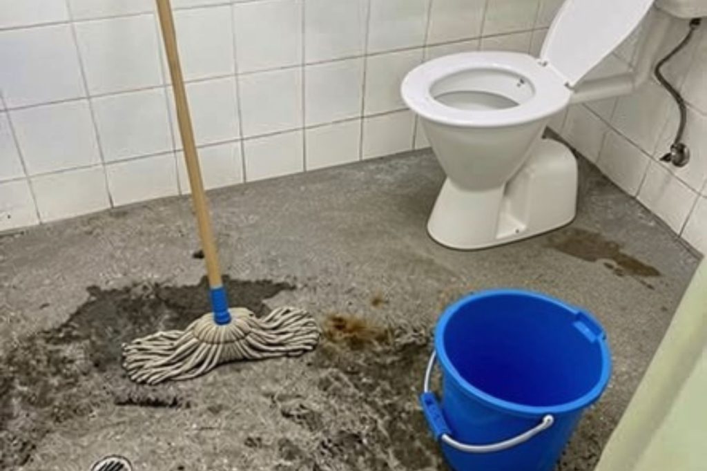 A mop and blue bucket are on a wet, dirty bathroom floor next to a white toilet with the lid open. The walls are tiled white.