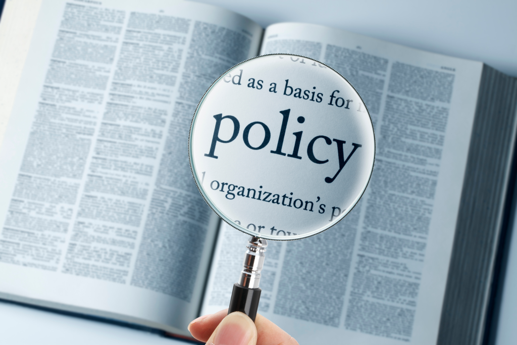 A hand holds a magnifying glass over an open dictionary, enlarging the word policy on the page.
