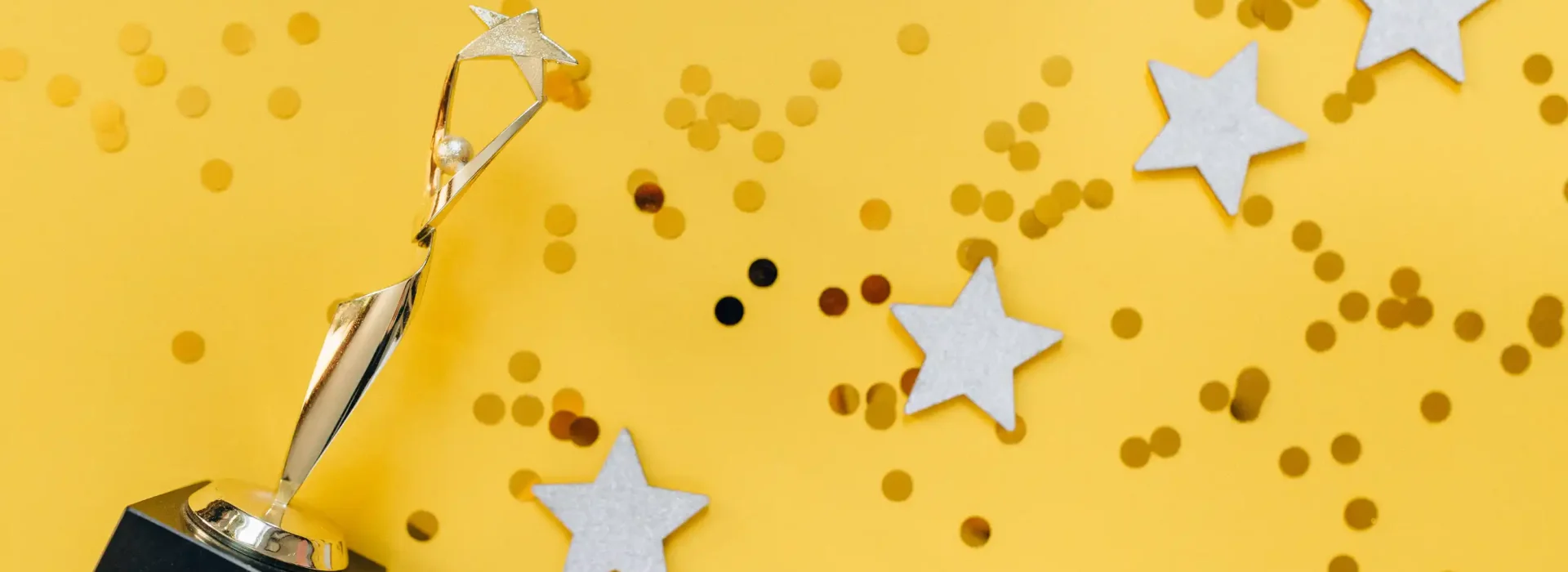 A gold trophy with a star stands on a yellow background, surrounded by gold confetti and five white star cutouts.
