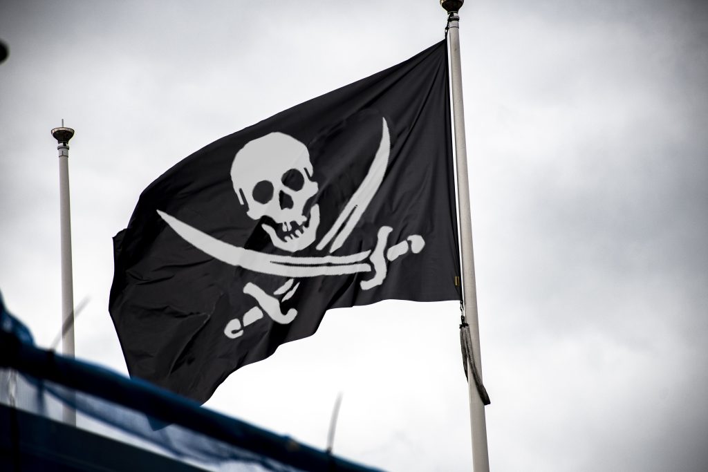 A black pirate flag with a white skull and crossbones above two crossed swords waves on a flagpole against a cloudy sky.