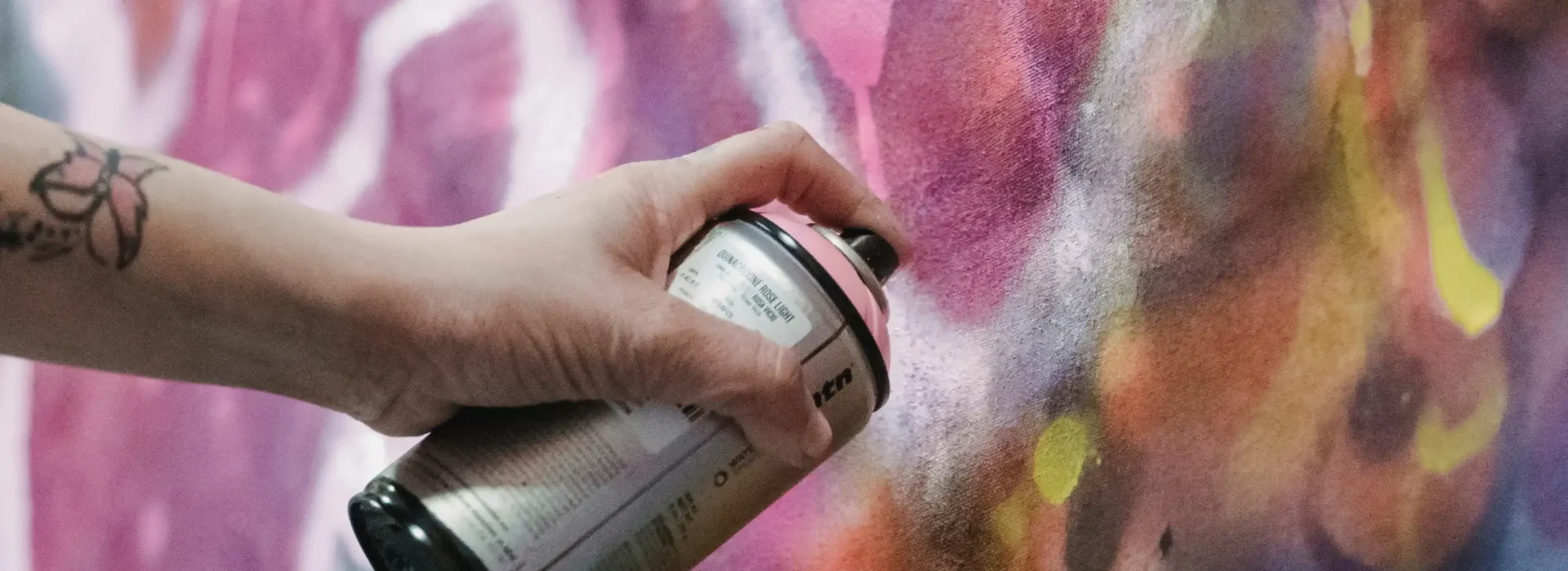A hand holding a spray paint can creates colorful graffiti art on a wall, with vivid pink, purple, orange, and yellow hues blending together.