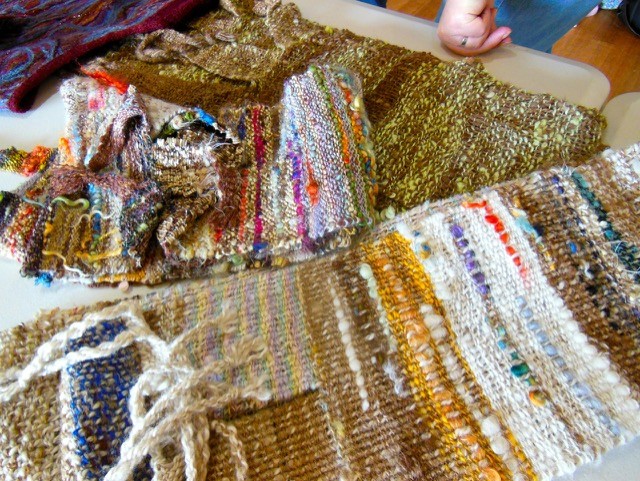 Handwoven textile pieces in earthy tones and colorful threads are spread on a table. A hand is visible in the top right corner, reaching toward the fabrics. The textiles have varied textures and patterns.