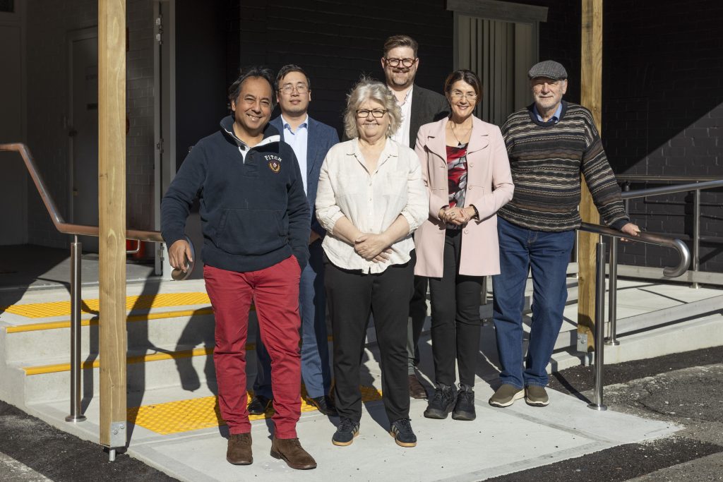 Six adults stand smiling outside a building entrance with steps and a ramp. They are casually dressed, and sunlight is shining on them. The background features a dark wall and a partially open door.