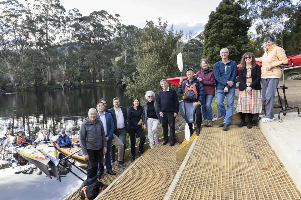 A group of people stands on a dock by the water, with kayaks and paddlers nearby. Trees and greenery are in the background, and a black dog is at the front.