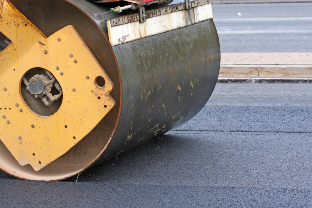 Close-up of a yellow steamrollers drum compacting fresh asphalt on a road, with part of the machine and smooth, dark pavement visible.