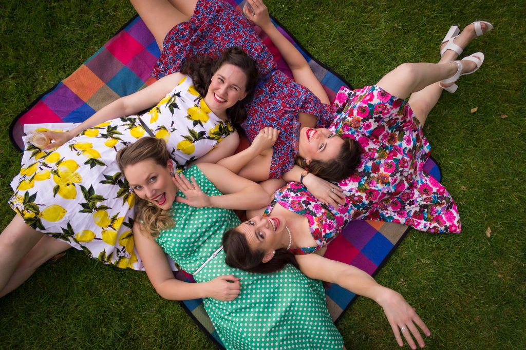 Four smiling women in colorful dresses lie on a picnic blanket on green grass, looking up at the camera. They appear happy and relaxed, enjoying time together outdoors.