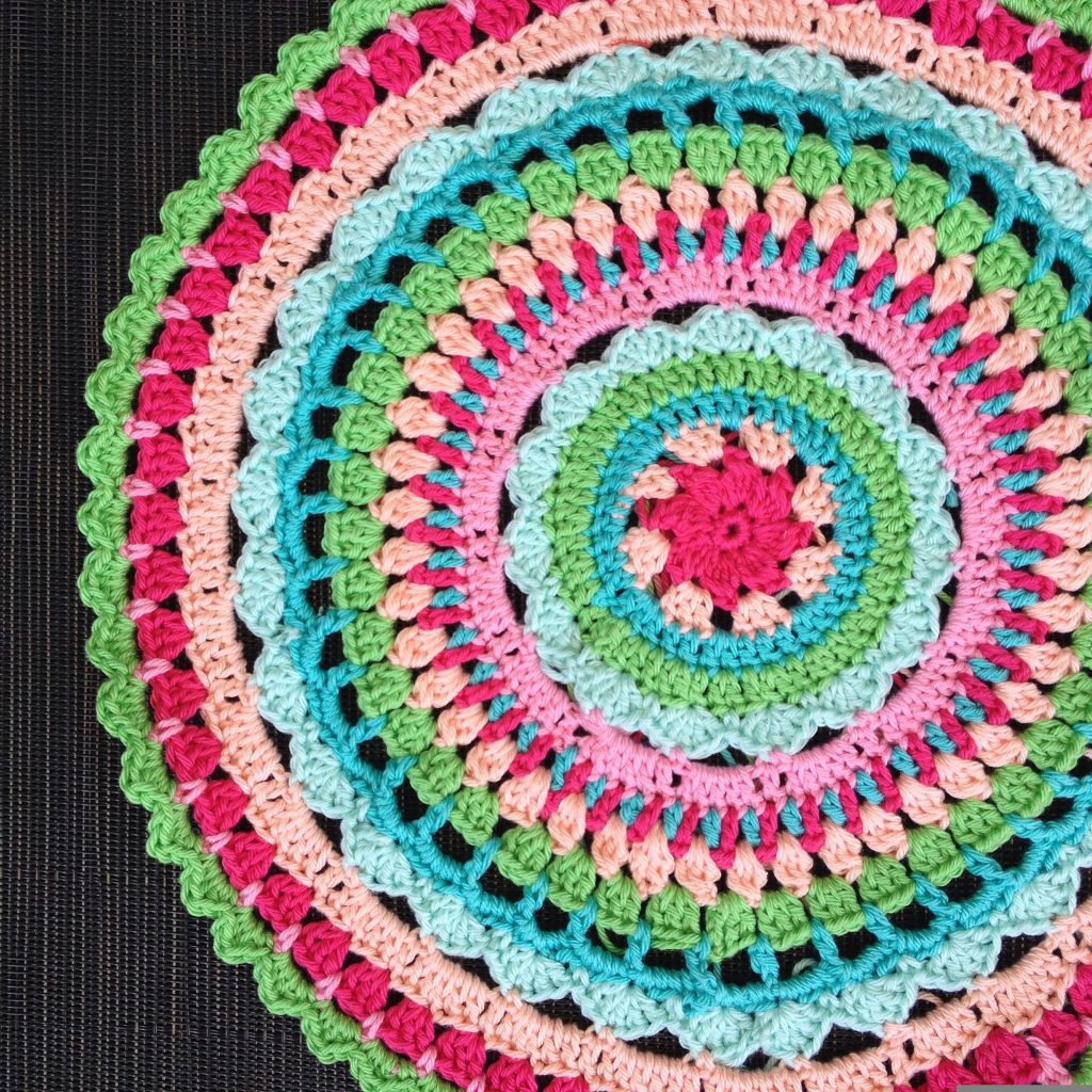 A colorful, circular crochet pattern featuring pink, green, blue, and peach yarn, displayed on a dark background. The rows have scalloped and textured designs.