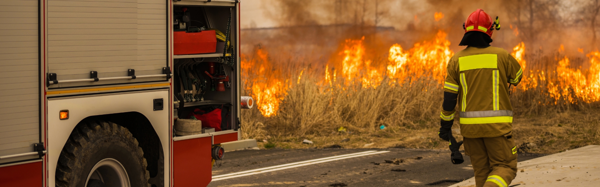 A firefighter in gear walks toward a grass fire near a road, while a fire truck with open compartments is parked nearby.