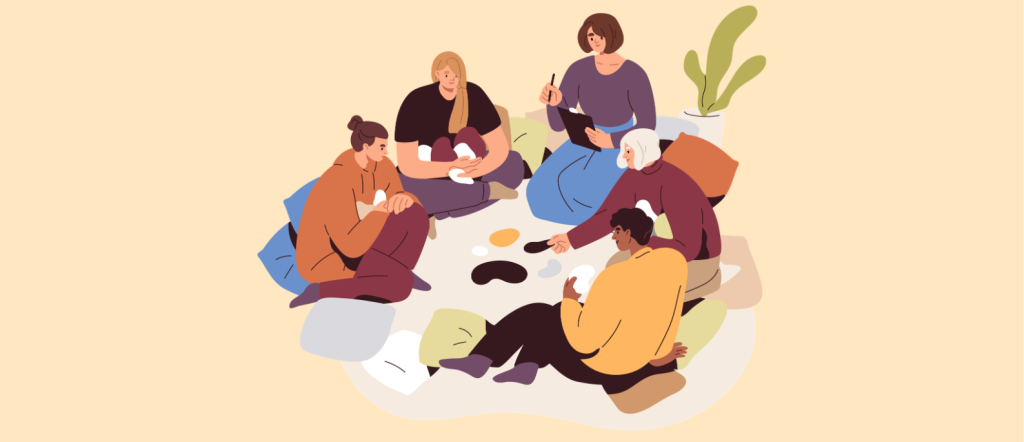 Five people sit together in a circle on the floor, talking and drinking from cups, surrounded by pillows and a plant, in a cozy, relaxed setting.