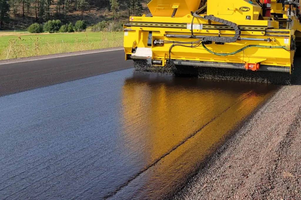 A yellow road paving machine applies a fresh layer of asphalt to a roadway, with smooth black pavement reflecting the machine in the sunlight.