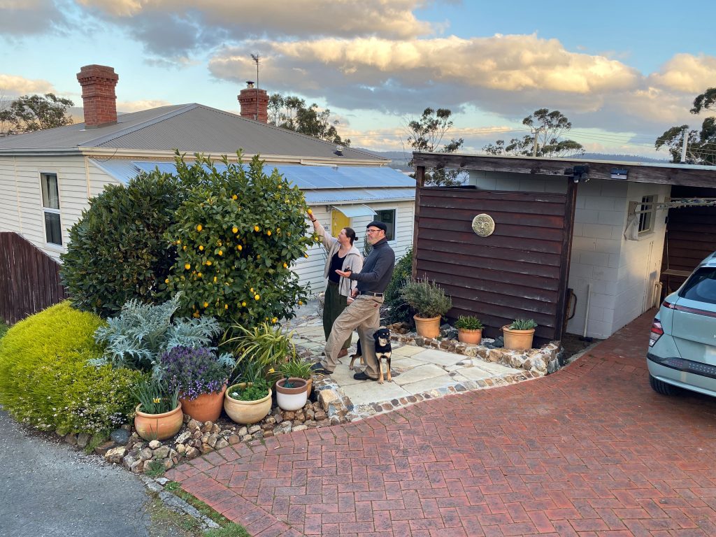 Two people and a dog stand on a stone path in front of a house with a brick driveway, surrounded by potted plants and flower bushes under a partly cloudy sky.