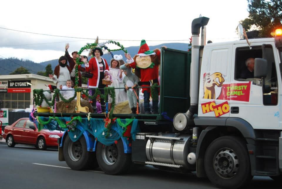 A white truck decorated for a holiday parade carries people in costumes, including an elf and an angel, on its flatbed.