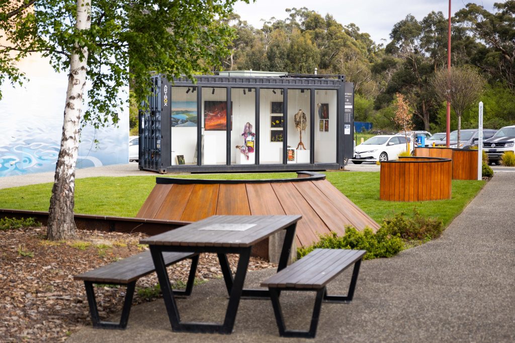 A shipping container converted into an art gallery display sits on a grassy area, with a picnic table in the foreground.