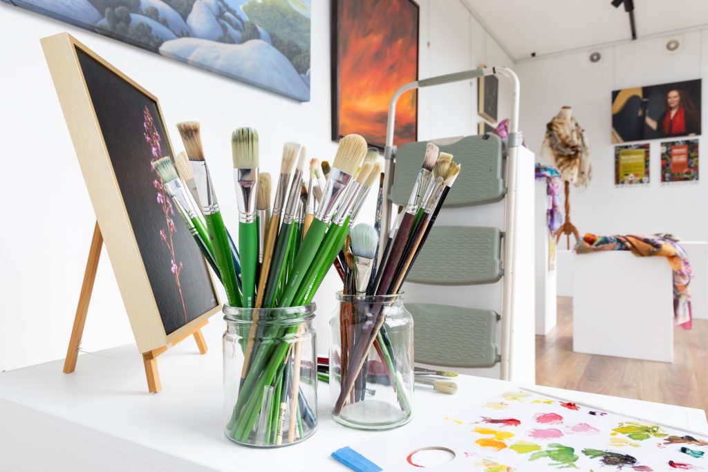Two glass jars filled with various paintbrushes sit on a white table next to a small framed painting and a palette of colorful paint dabs.