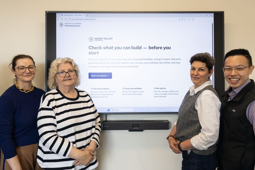 Four people standing in front of a large monitor displaying the Huon Valley Council planning enquiry website.