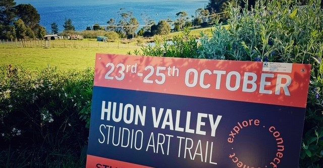 A lamb stands in a grassy field in front of a sign for the Huon Valley Studio Art Trail, held October 23rd to 25th, with a lake and hills in the background.