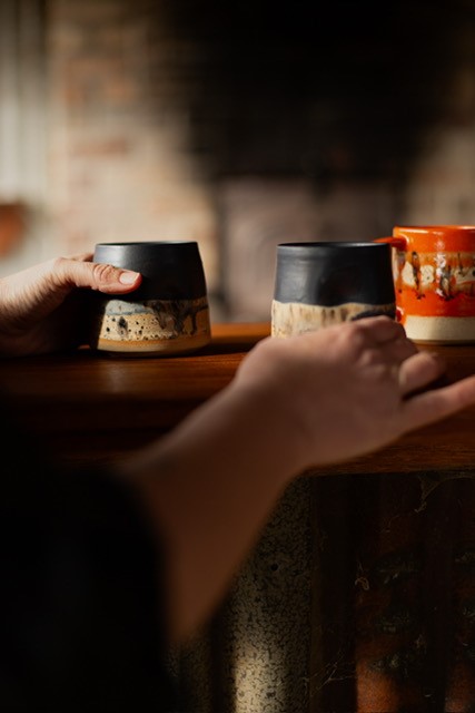 Hands place two ceramic mugs with black rims and textured bases on a wooden surface next to an orange mug.