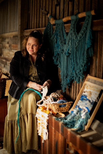 A woman in a long dress and jacket sits in a studio, working on a fiber art project with weaving tools and yarn nearby.