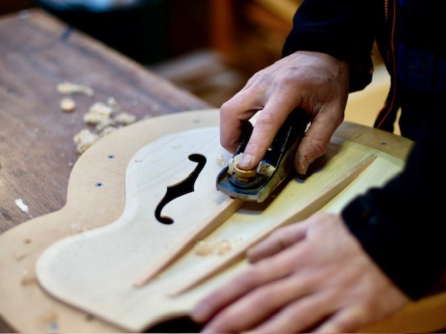 A woodworker uses a small hand plane to shape the top of an unfinished acoustic guitar.