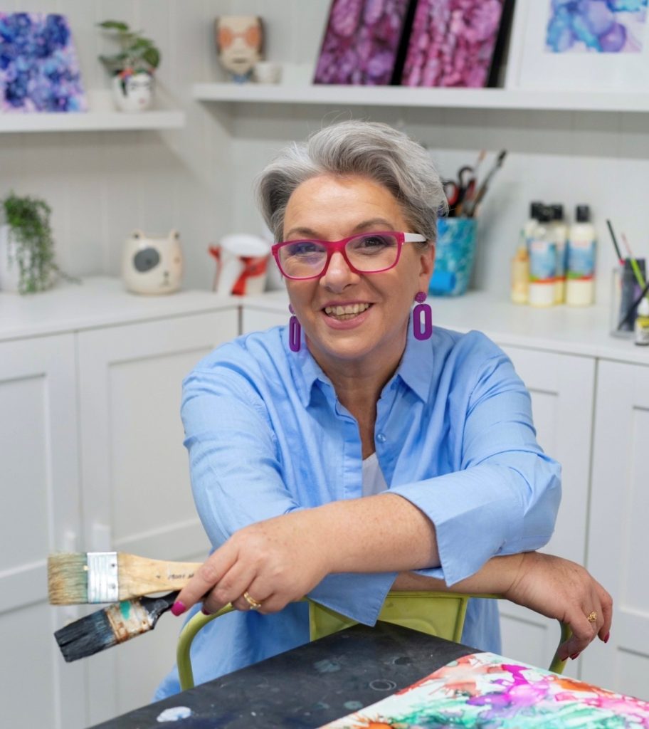 A woman with short gray hair and bright pink glasses holds two paintbrushes while smiling in her art studio.