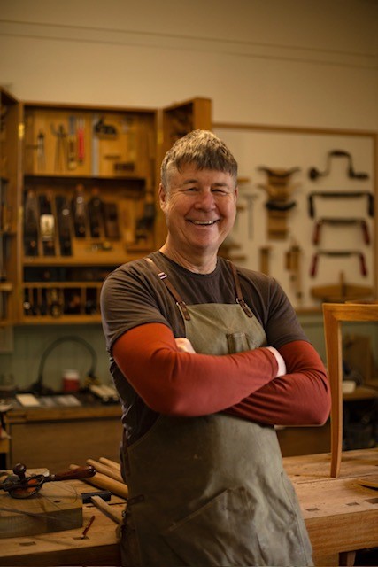 A smiling woodworker in an apron stands with his arms crossed in a workshop filled with hand tools.