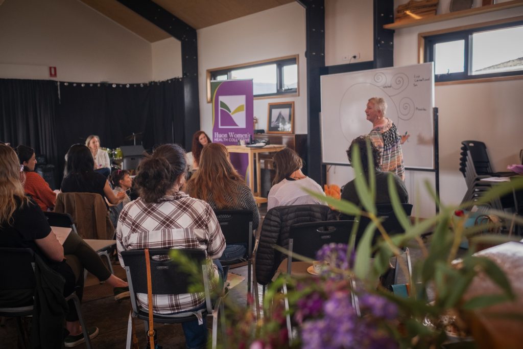 A woman stands at a whiteboard leading a discussion for a group of attendees seated in a workshop space.