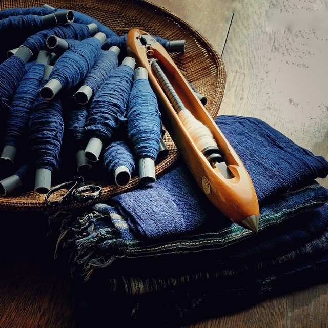 A wooden weaving shuttle resting on a stack of blue textiles next to a basket filled with bobbins of blue thread.