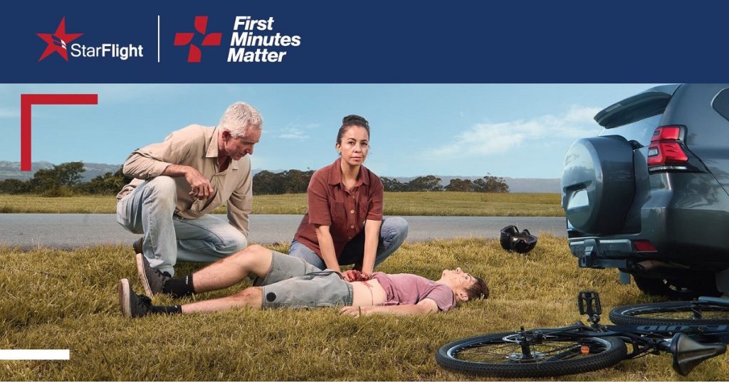 Two people kneeling on grass perform first aid on a cyclist lying on the ground next to a bicycle and a parked car. The StarFlight and First Minutes Matter logos are at the top.