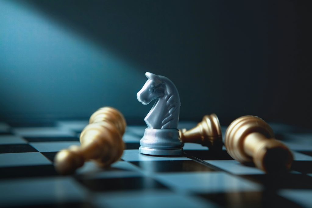 A silver chess knight standing upright on a chessboard surrounded by several fallen gold chess pieces.