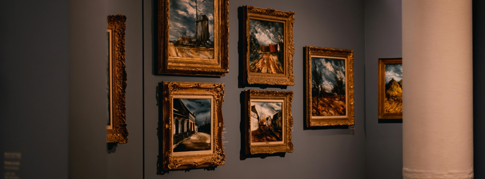 Several framed oil paintings of landscapes and street scenes displayed on a dark gallery wall.